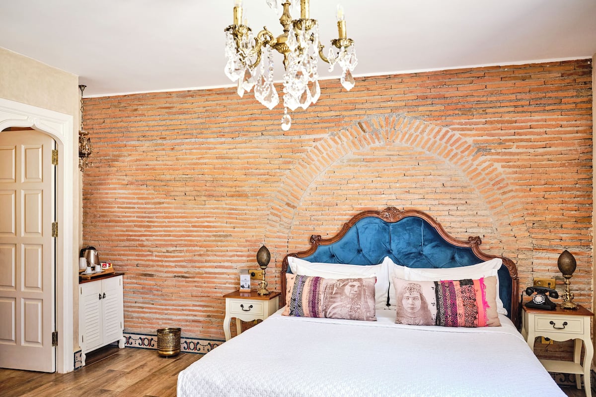 A bedroom with a brick wall, a chandelier, a bed with blue headboard, and a nightstand with a telephone.
