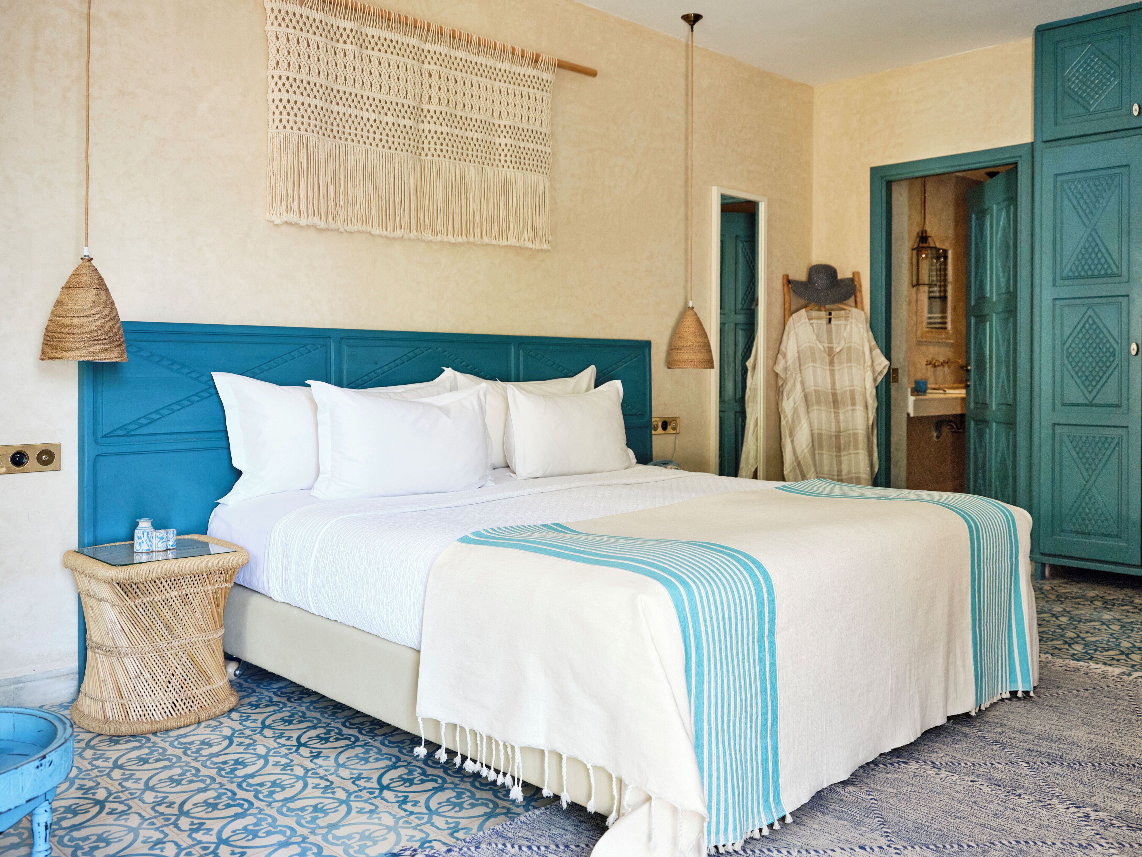 Una cama con ropa de cama blanca, un cabecero azul y una manta con rayas azules. Encima de la cama hay un tapiz tejido y una mesita de noche de mimbre con un cuenco azul pequeño a su lado.
