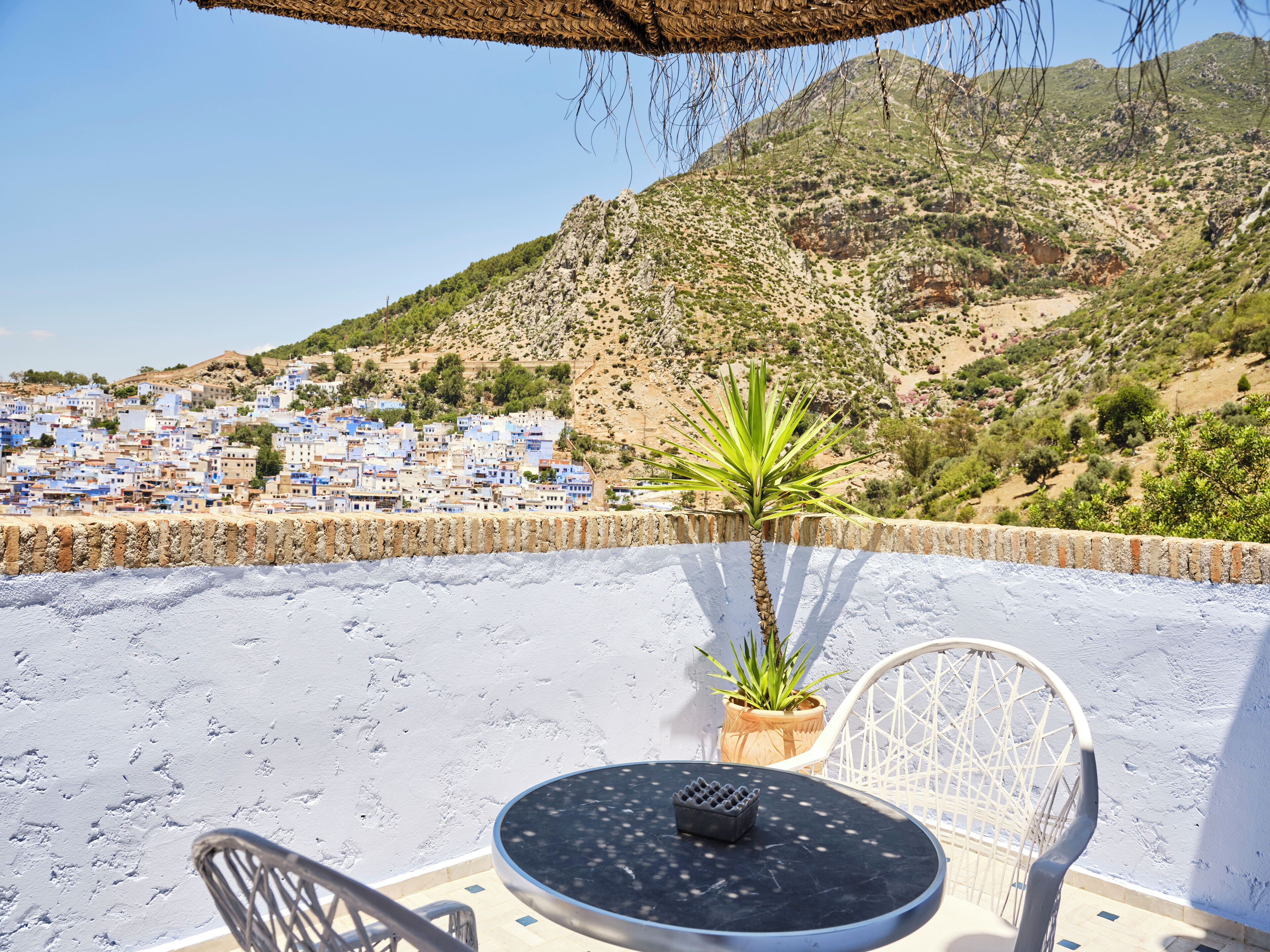 Una terraza con una mesa y sillas, con vistas a un pueblo costero de edificios azules y montañas al fondo.