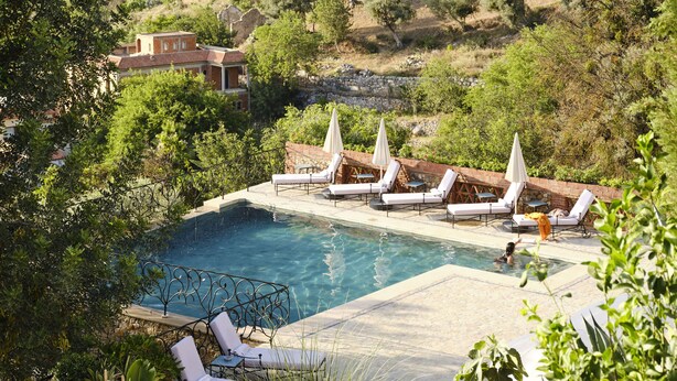 Un área de piscina con sillones de descanso y sombrillas, rodeada de vegetación y con montañas al fondo.
