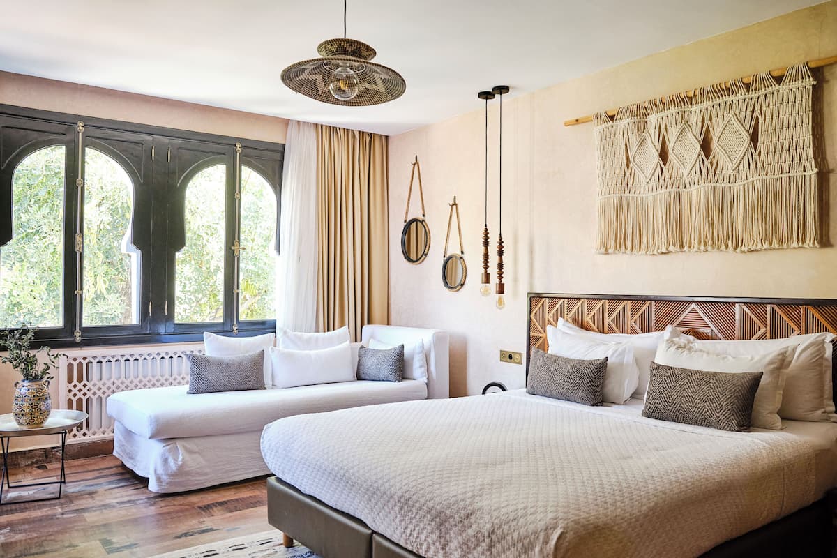 A bedroom with two beds, a large window with curtains, a macrame wall hanging, and a ceiling light fixture.