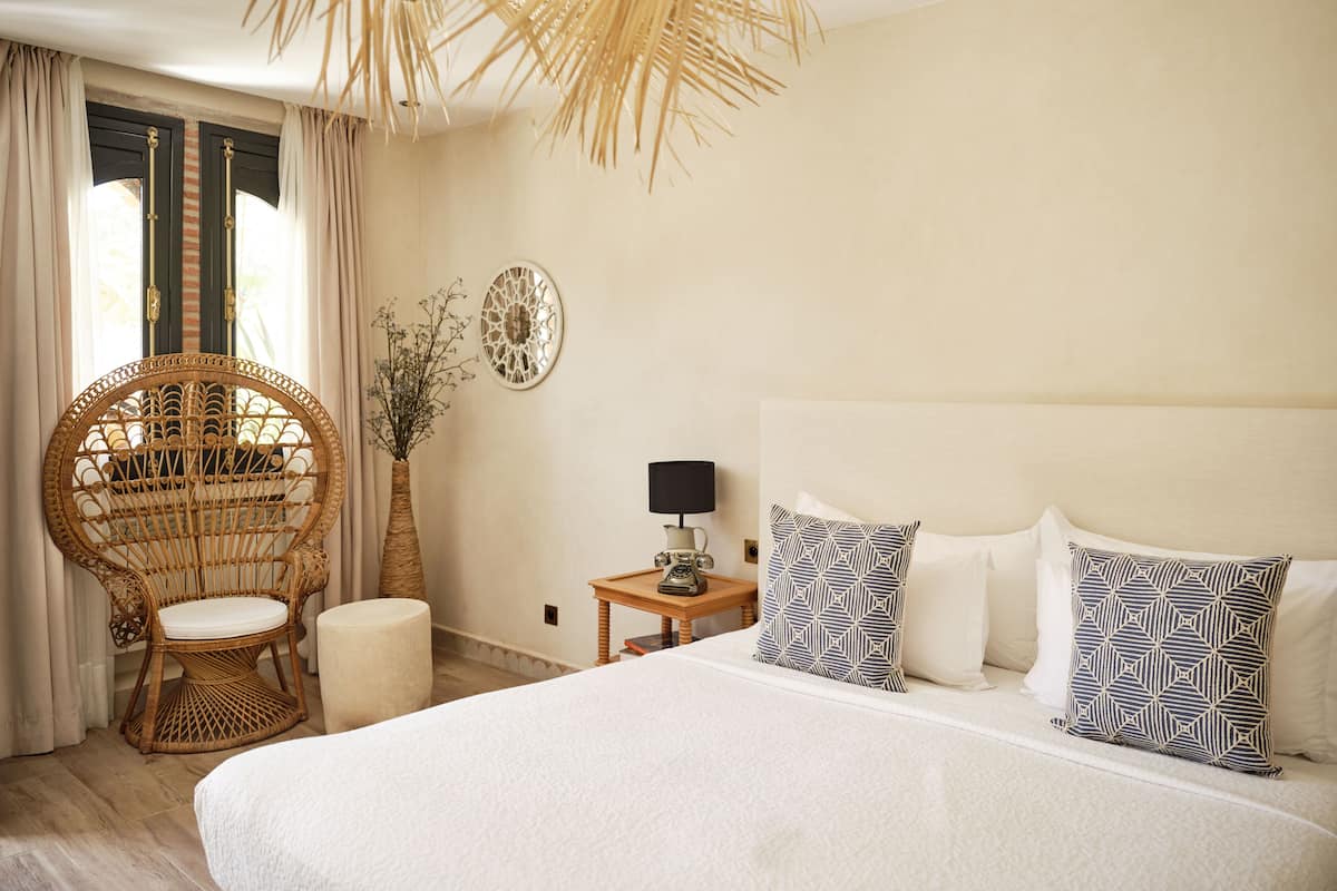 A spacious bedroom with a large bed, a wicker chair, a small bedside table with a lamp, and a decorative wall piece.