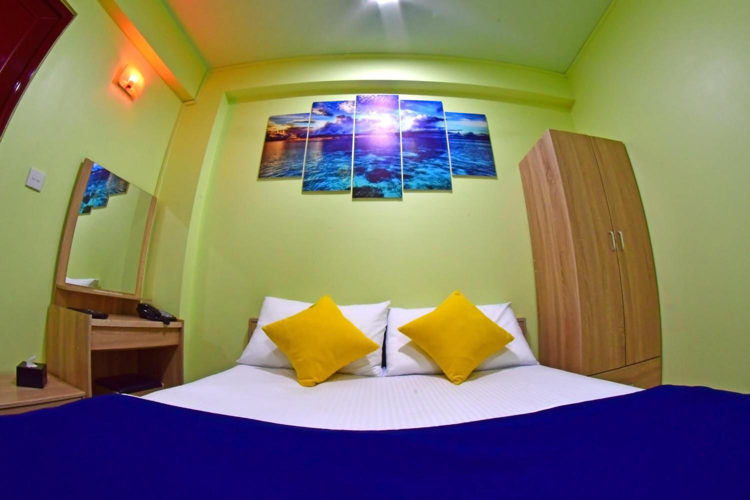 A hotel room with a bed, two yellow pillows, a wooden wardrobe, a nightstand with a phone, and a wall-mounted painting.