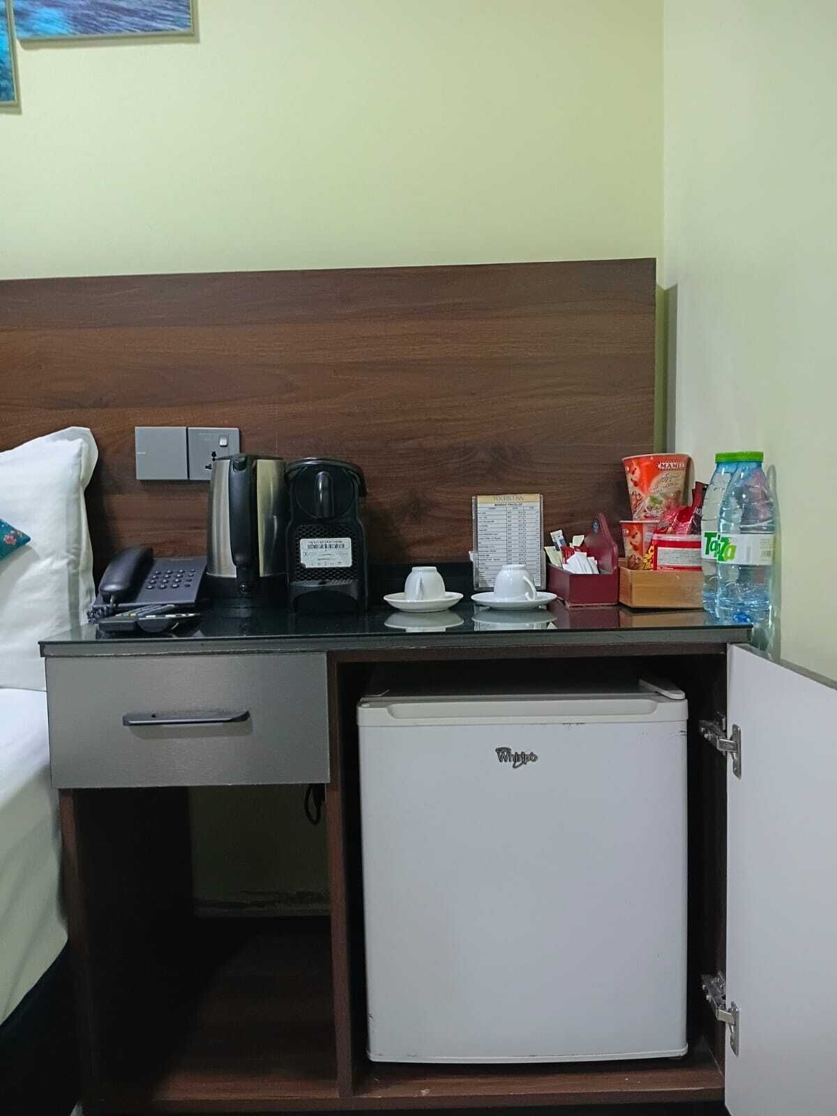 Una habitación de hotel con cama, escritorio, teléfono, cafetera, refrigerador pequeño y algunos alimentos.
