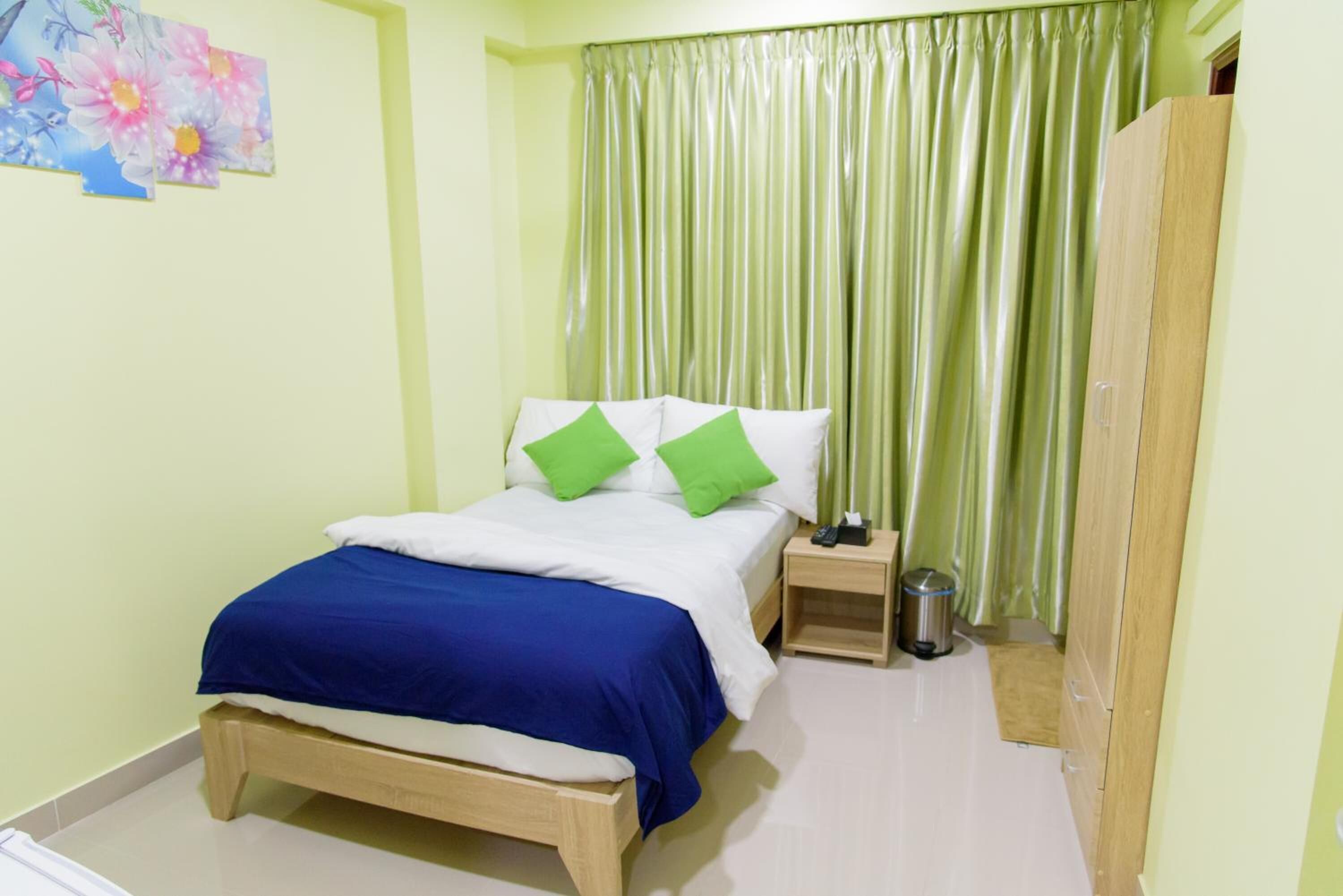 A bedroom with a bed, green pillows, a nightstand, a trash bin, and a wardrobe.