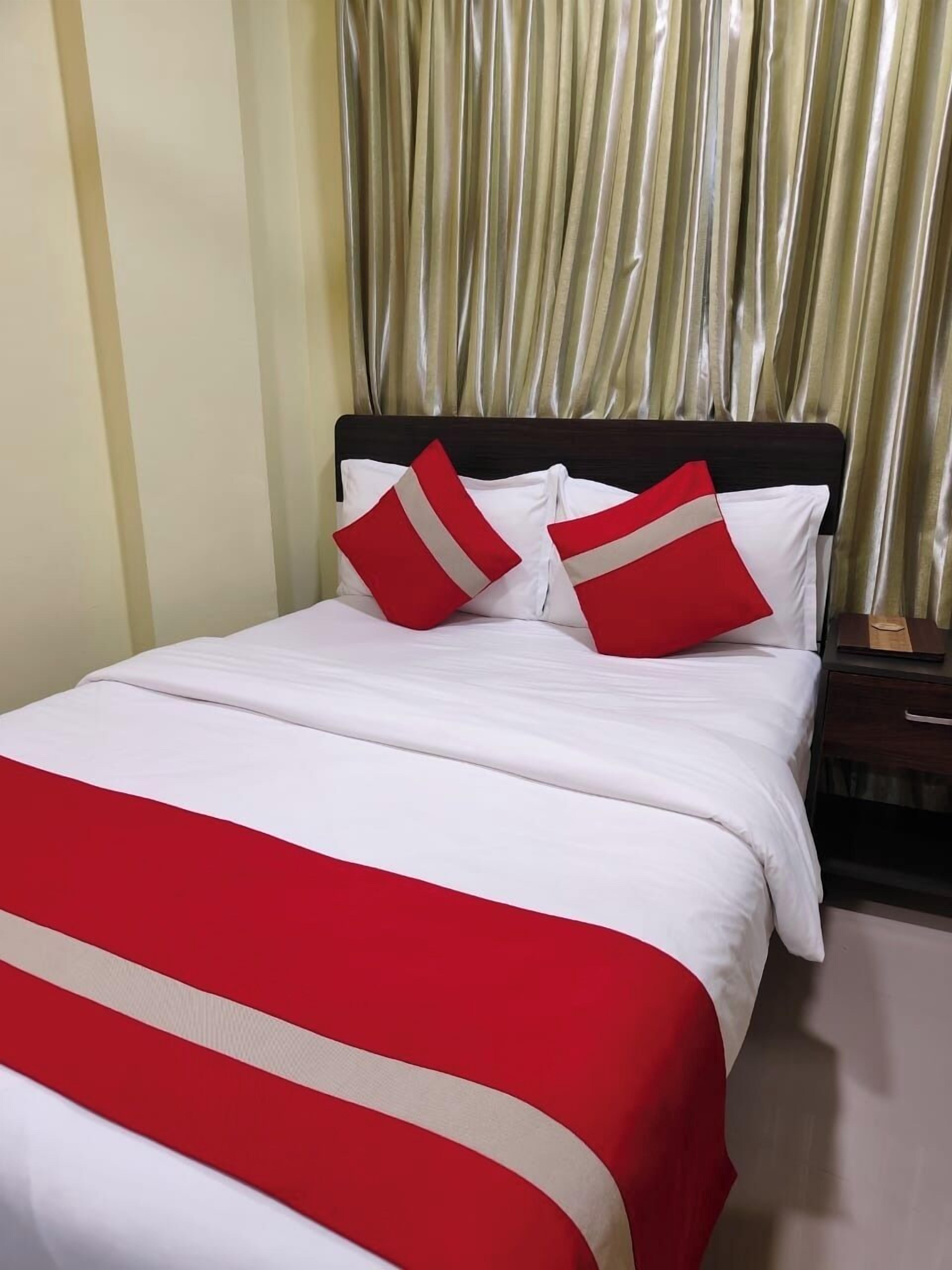 Una cama con ropa de cama blanca y almohadas rojas y blancas, un cabecero de madera y una cortina dorada al fondo.