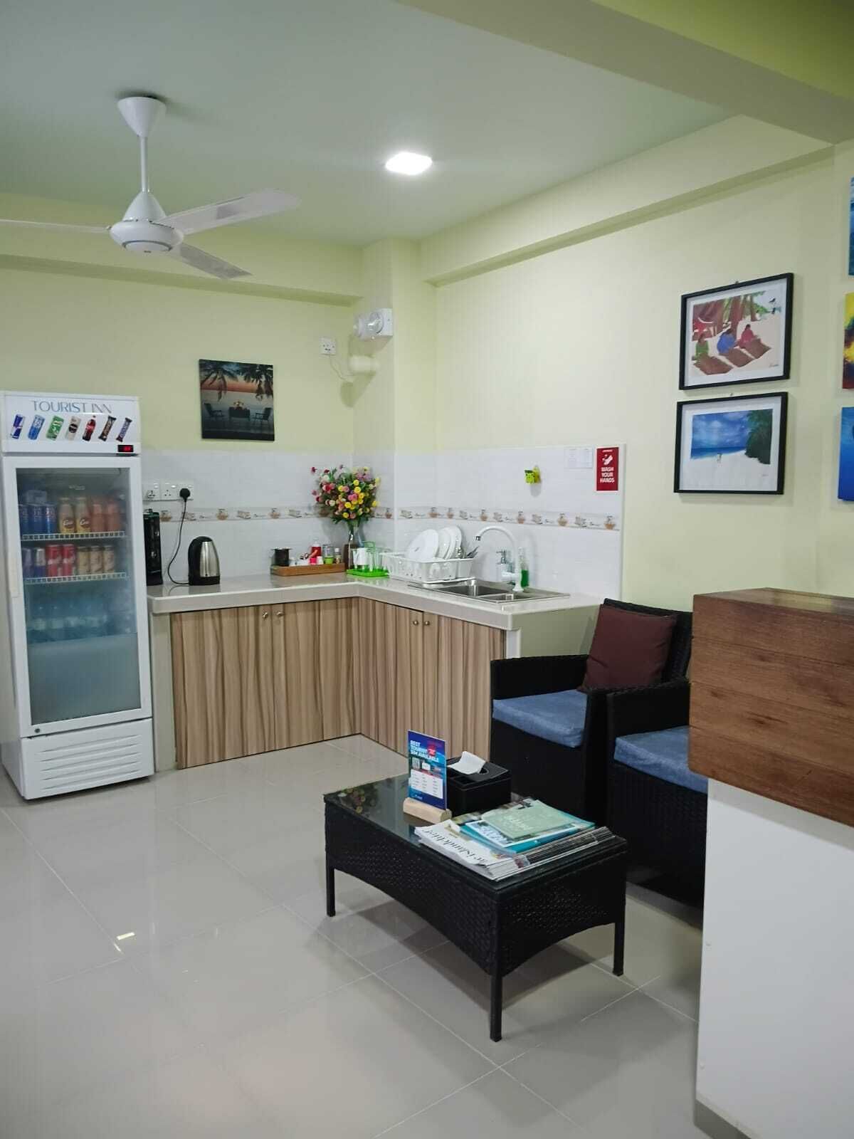 A hotel lobby with a kitchenette, a refrigerator, a coffee machine, a flower vase, and framed pictures on the wall.
