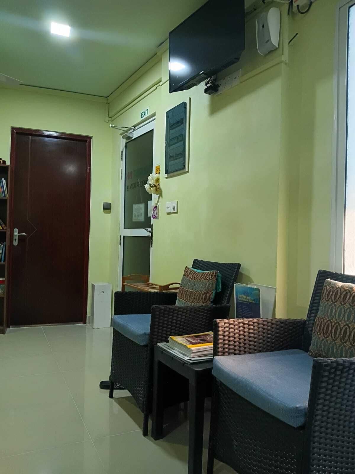 Um quarto com uma porta marrom escura, parede branca, TV de tela plana acima de uma janela e uma pequena mesa com uma cadeira.