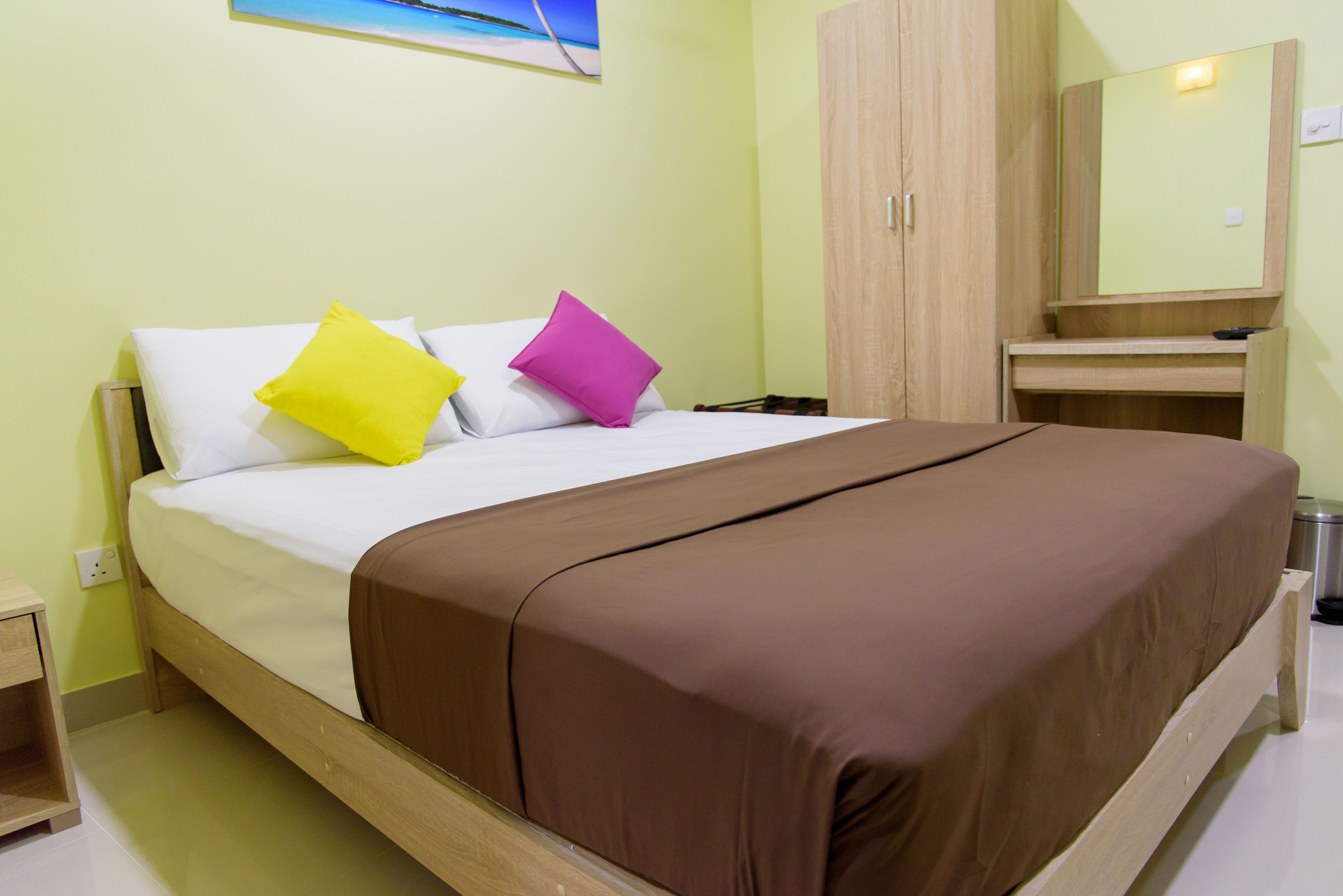 Una cama con ropa de cama blanca y marrón, dos almohadas en amarillo y morado, un cabecero de madera, una mesita de noche, un armario de madera y un espejo sobre el armario.