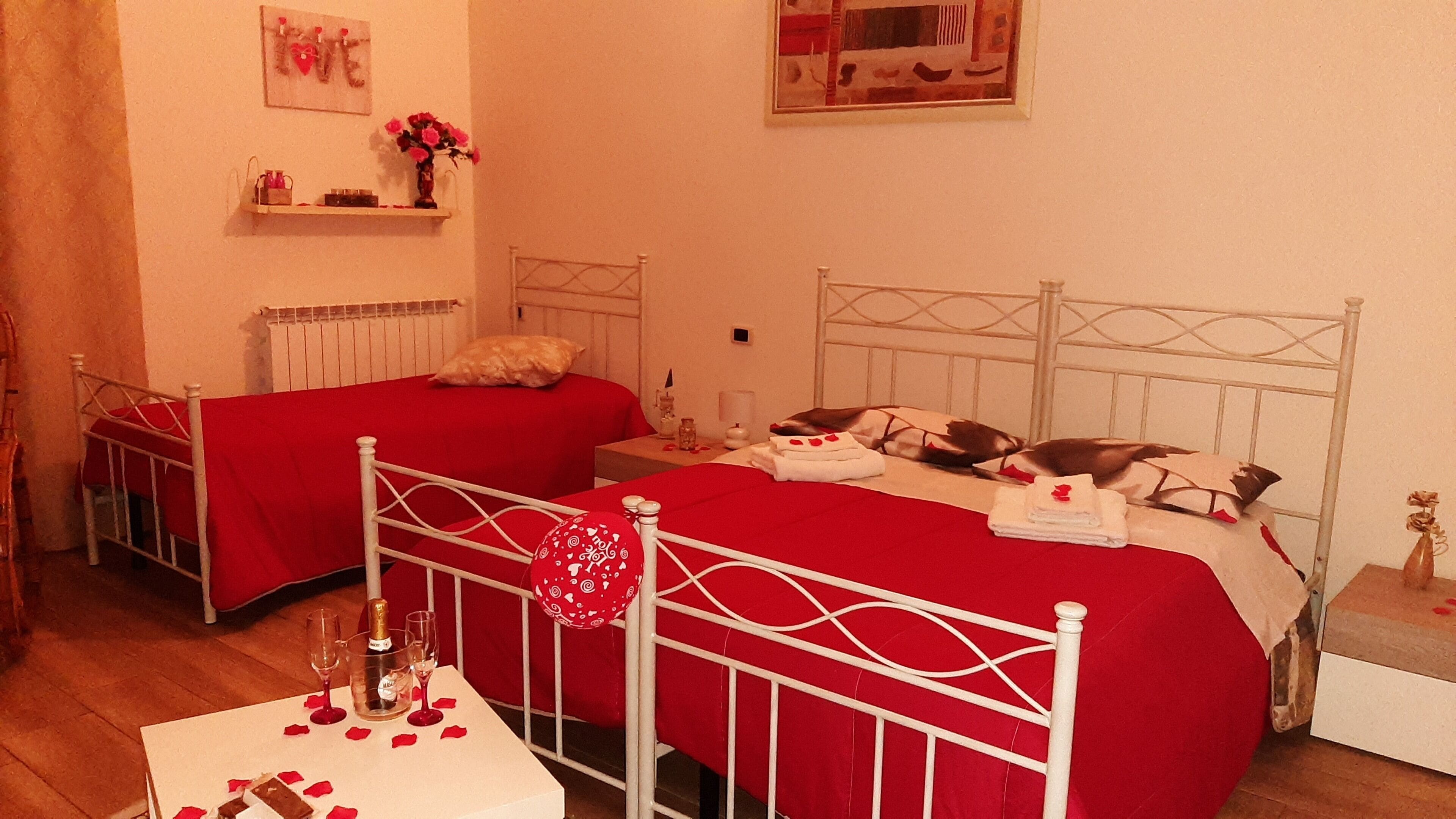 Camera d'albergo con due letti, ciascuno con copriletto rosso e testata bianca in metallo. C'è un tavolino con una bottiglia di champagne e bicchieri, e un palloncino rosso sul pavimento.