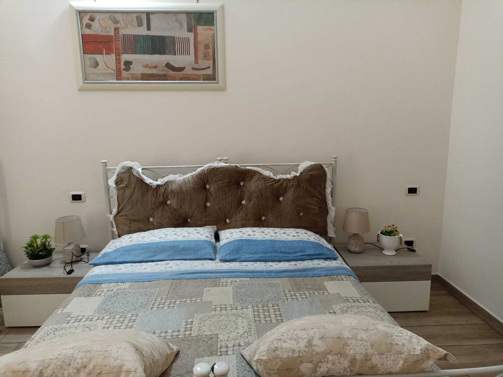 A bedroom with a bed, two bedside tables, a wall-mounted lamp, and a small potted plant.