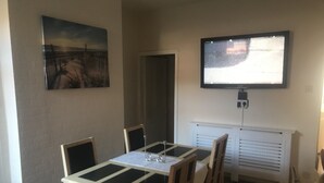 A dining area with a table and chairs, a painting on the wall, and a television mounted above a white radiator. - SOUTH LAKES TOWN CENTRE HOUSE (Barrow-in-Furness)