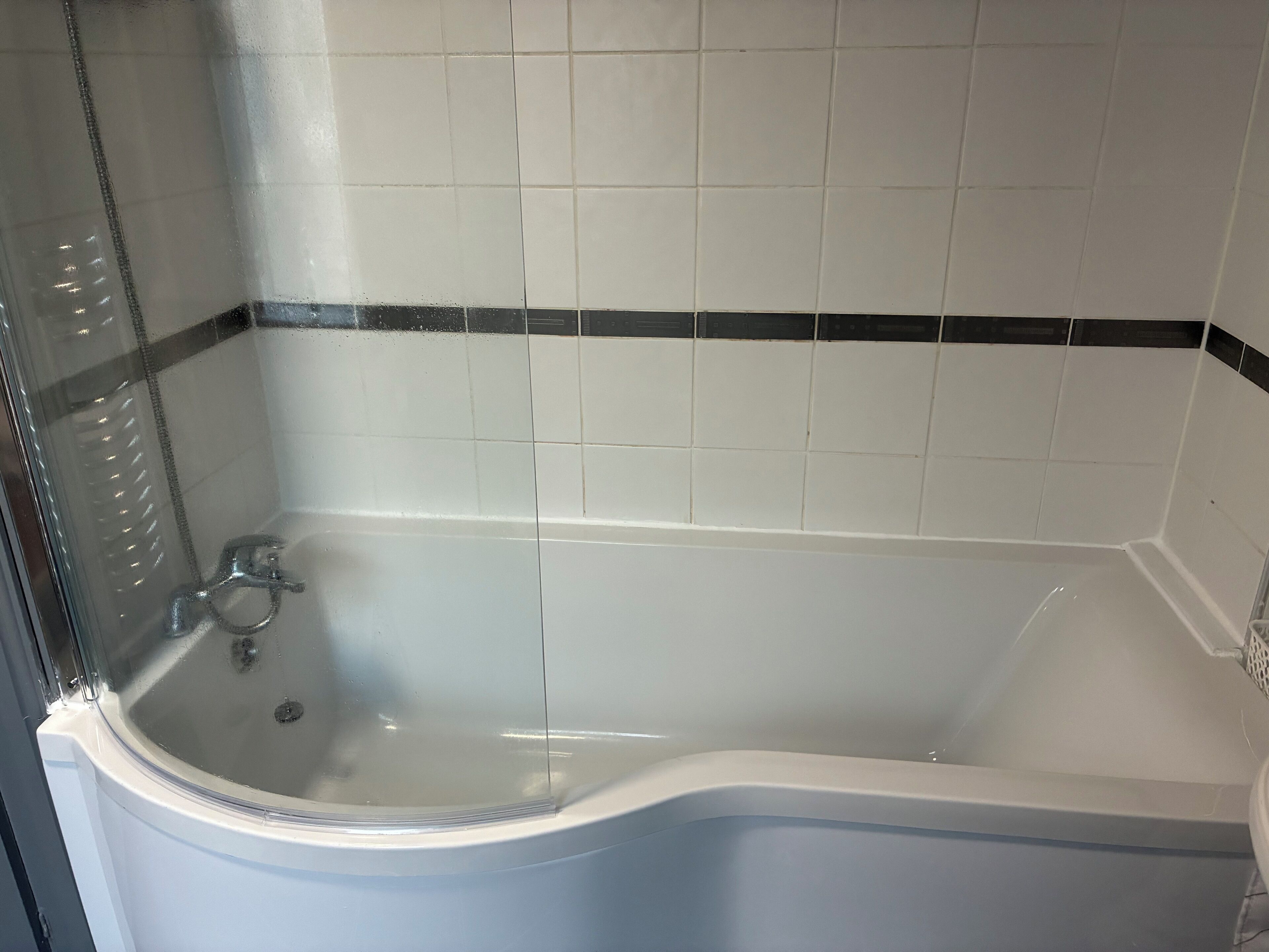 Una bañera con cerramiento de vidrio, azulejos blancos con borde negro y desagüe visible.