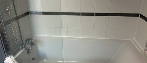 Una bañera con cerramiento de vidrio, azulejos blancos con borde negro y desagüe visible.