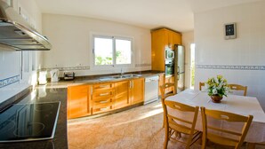 Een keuken met een fornuis, spoelbak en kasten, een eettafel met stoelen en een raam dat voor natuurlijk licht zorgt.