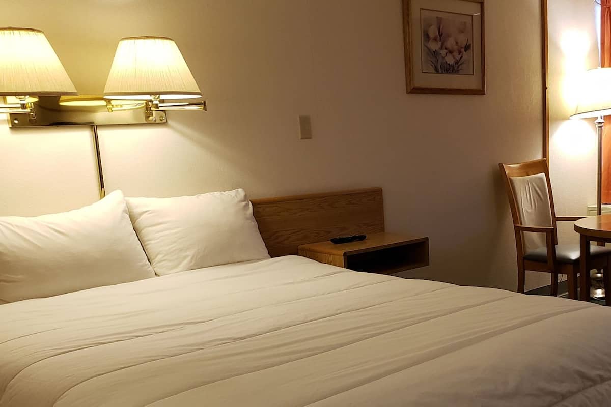 A hotel room with a bed, two bedside lamps, a chair, a small table, and a window with curtains.