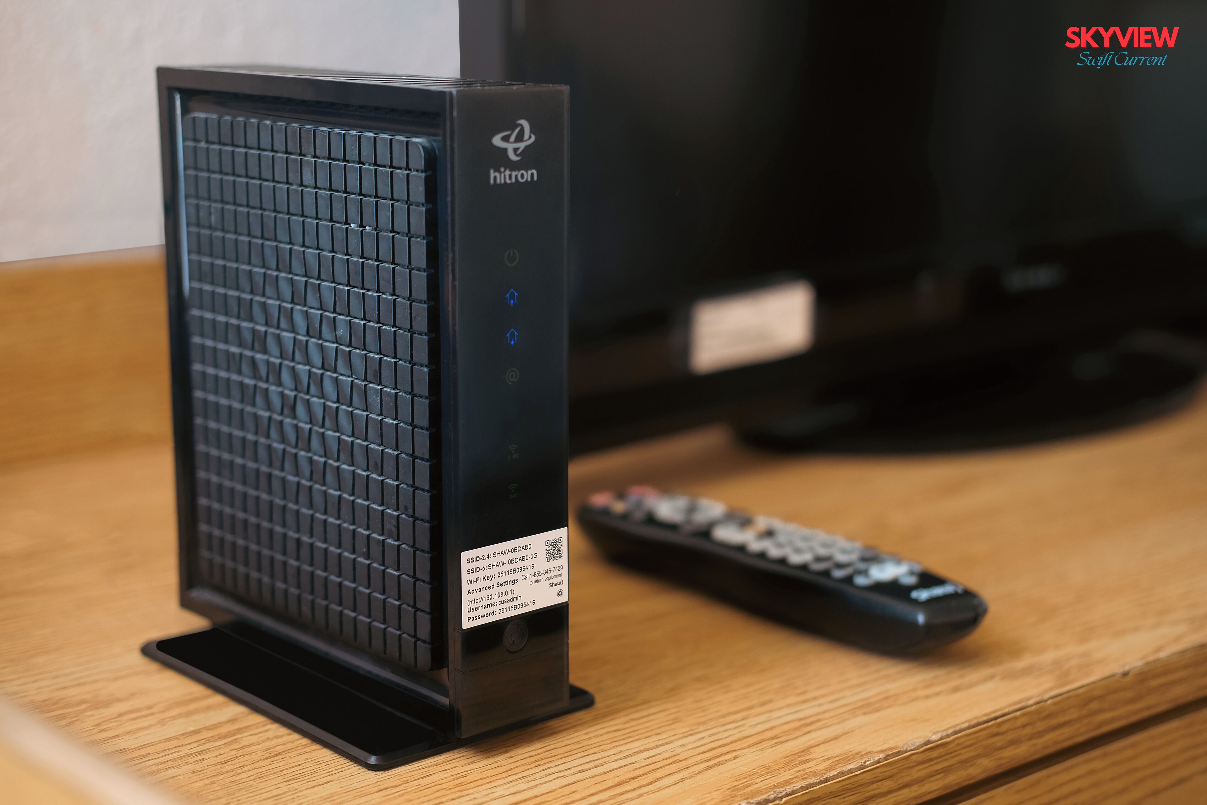 Un appareil électronique noir, doté d’un motif quadrillé sur la façade, une télécommande et un téléviseur en arrière-plan.