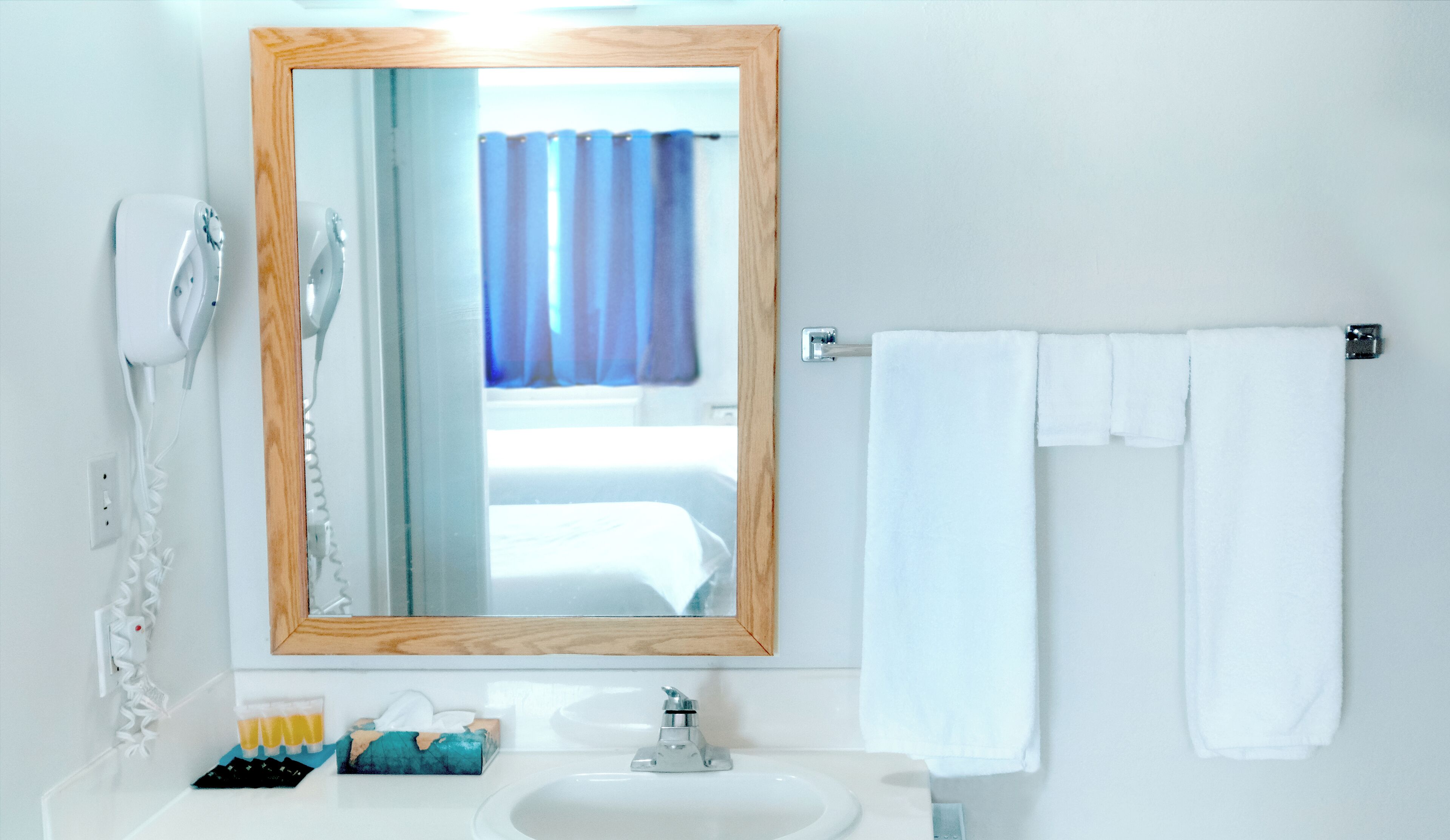 Ein Badezimmer mit einem spiegelnden Holzelement, einem weißen Waschbecken, einem Handtuchhalter mit zwei weißen Handtüchern, einem Fön und einem Fenster mit blauen Vorhängen.