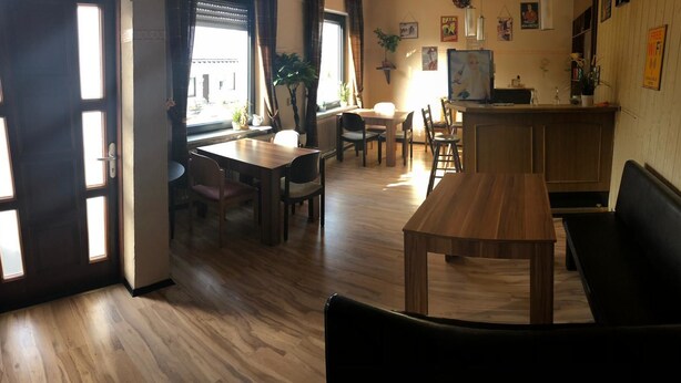 A dining area with wooden flooring, a central table, and chairs. There is a bar counter with a refrigerator and a potted plant. - Pension Lume (Oberraden)