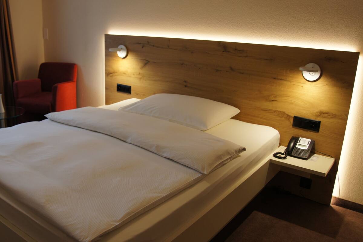 Een hotelkamer met een bed, een rode stoel, een nachtkastje met een telefoon en een houten hoofdeinde.
