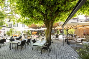 An outdoor café with tables and chairs arranged under a large tree, surrounded by buildings and potted plants. - Ochsen Lenzburg (Lenzburg)