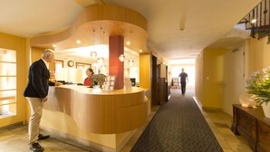A hotel reception area with a curved wooden counter, a receptionist, and a man standing by the counter.