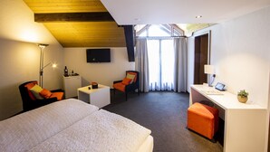 A hotel room with a bed, a desk, a chair, a small table, a TV, and a window with curtains.