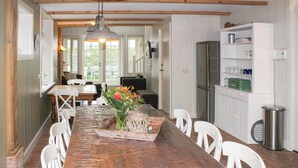 Een keuken met een houten tafel, witte stoelen, een koelkast en een wit kastje met planken.