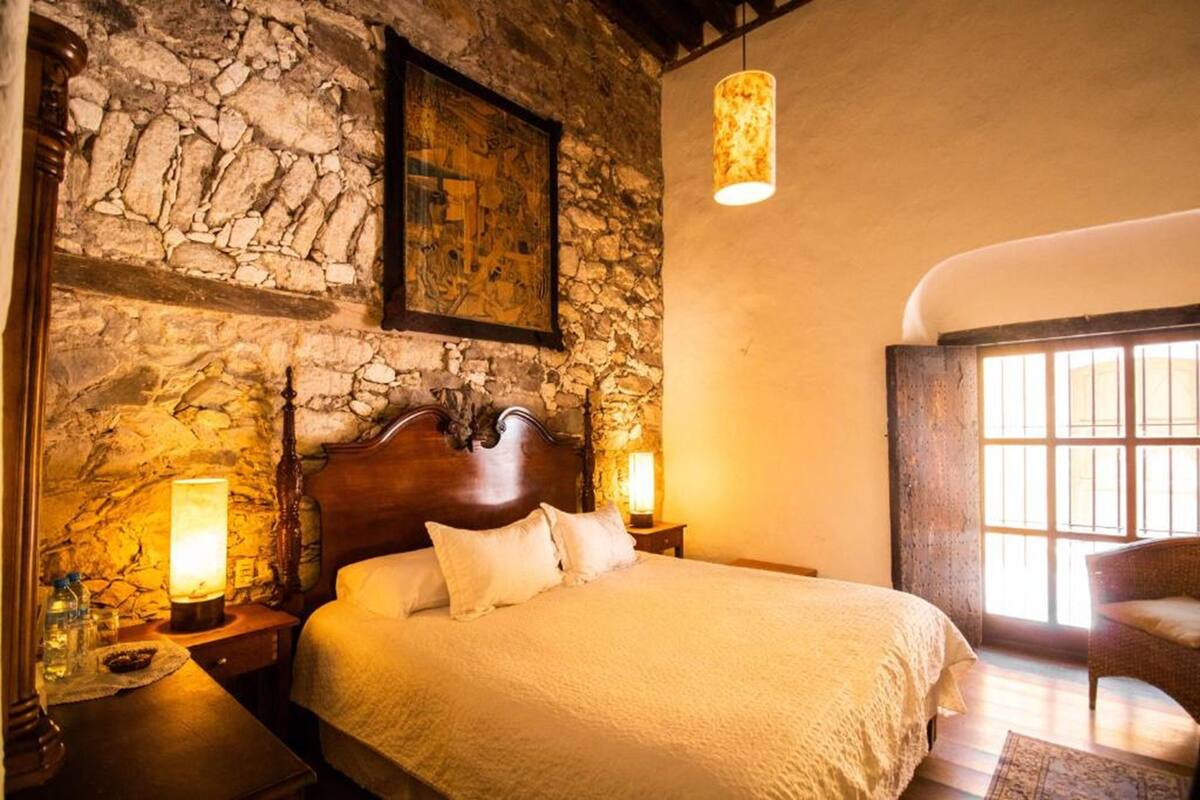 Une chambre avec un mur en pierre, un lit en bois, deux lampes de chevet et une fenêtre laissant entrer la lumière naturelle.