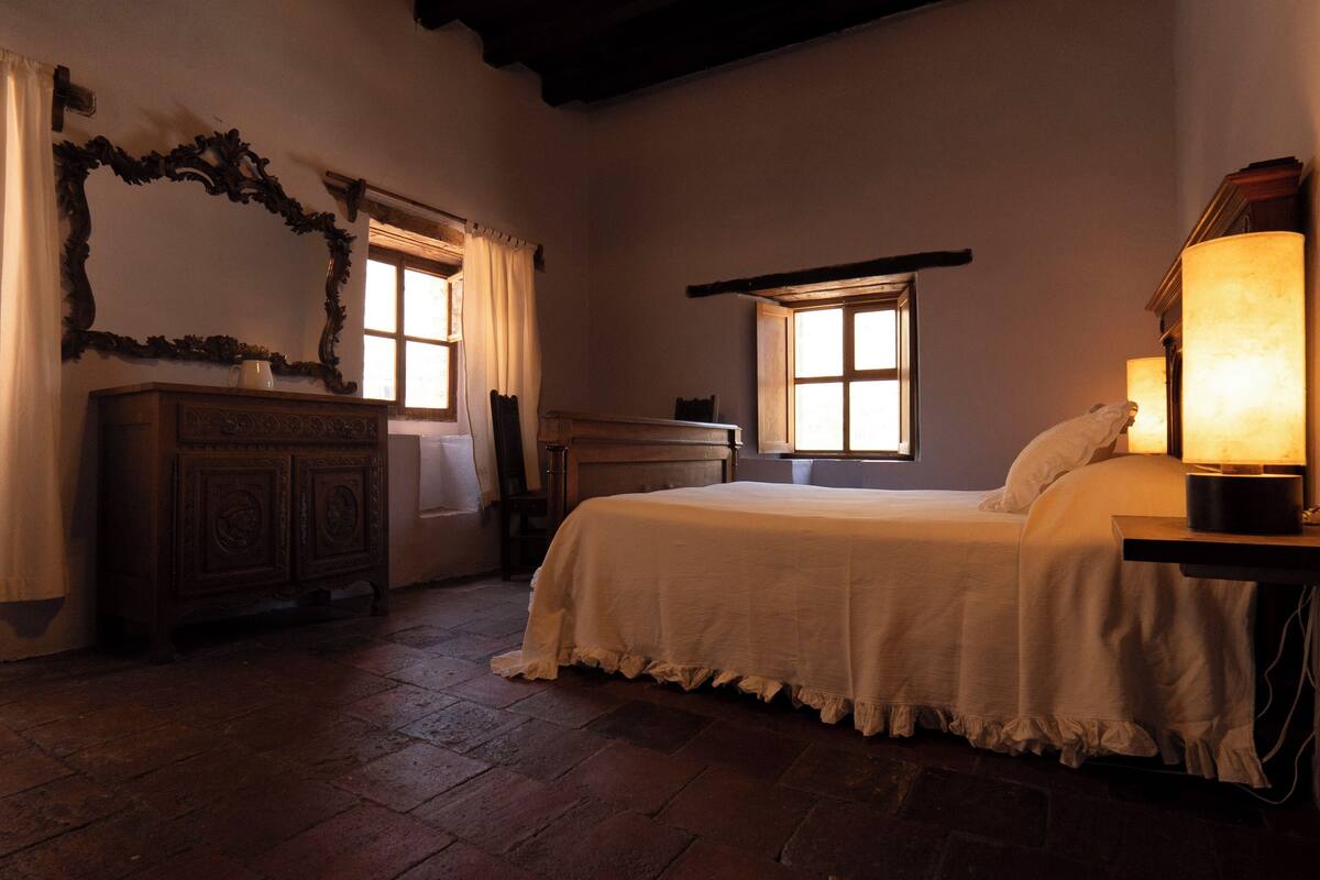 Une pièce comprenant un lit, une commode, deux fenêtres et une lampe.