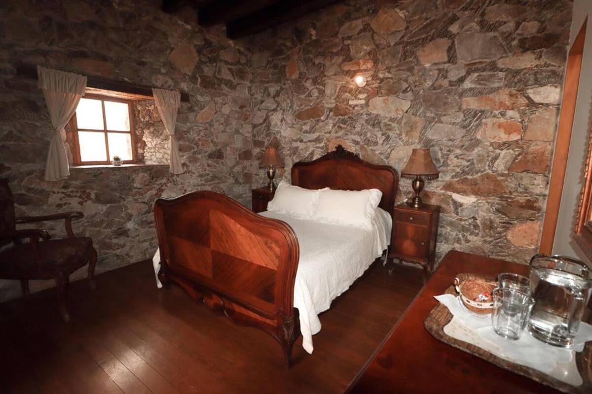 Une chambre aux murs de pierre, avec un lit en bois, une table de chevet avec une lampe et une petite table sur laquelle se trouvent des verres et des en-cas.