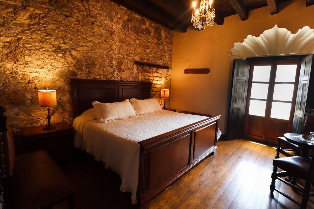 Une chambre avec un mur en pierre, un lit en bois, deux lampes de chevet, une chaise en bois et un lustre.