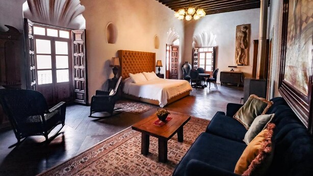 Habitación amplia con una cama grande, un sofá, una mesa de centro y una silla. Cuenta con una ventana con cortinas, un candelabro y un cuadro en la pared.