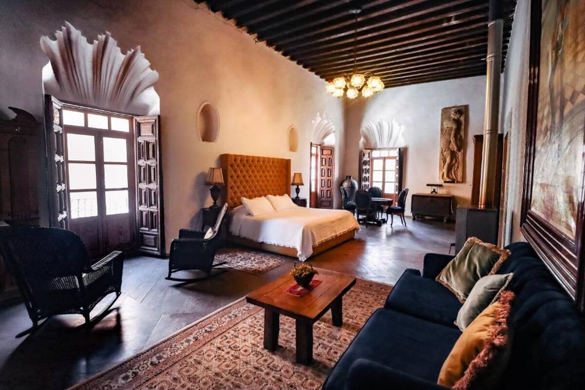 Une pièce spacieuse comprenant un grand lit, un canapé, une table basse et un fauteuil. On y trouve une fenêtre avec des rideaux, un lustre et un tableau accroché au mur.