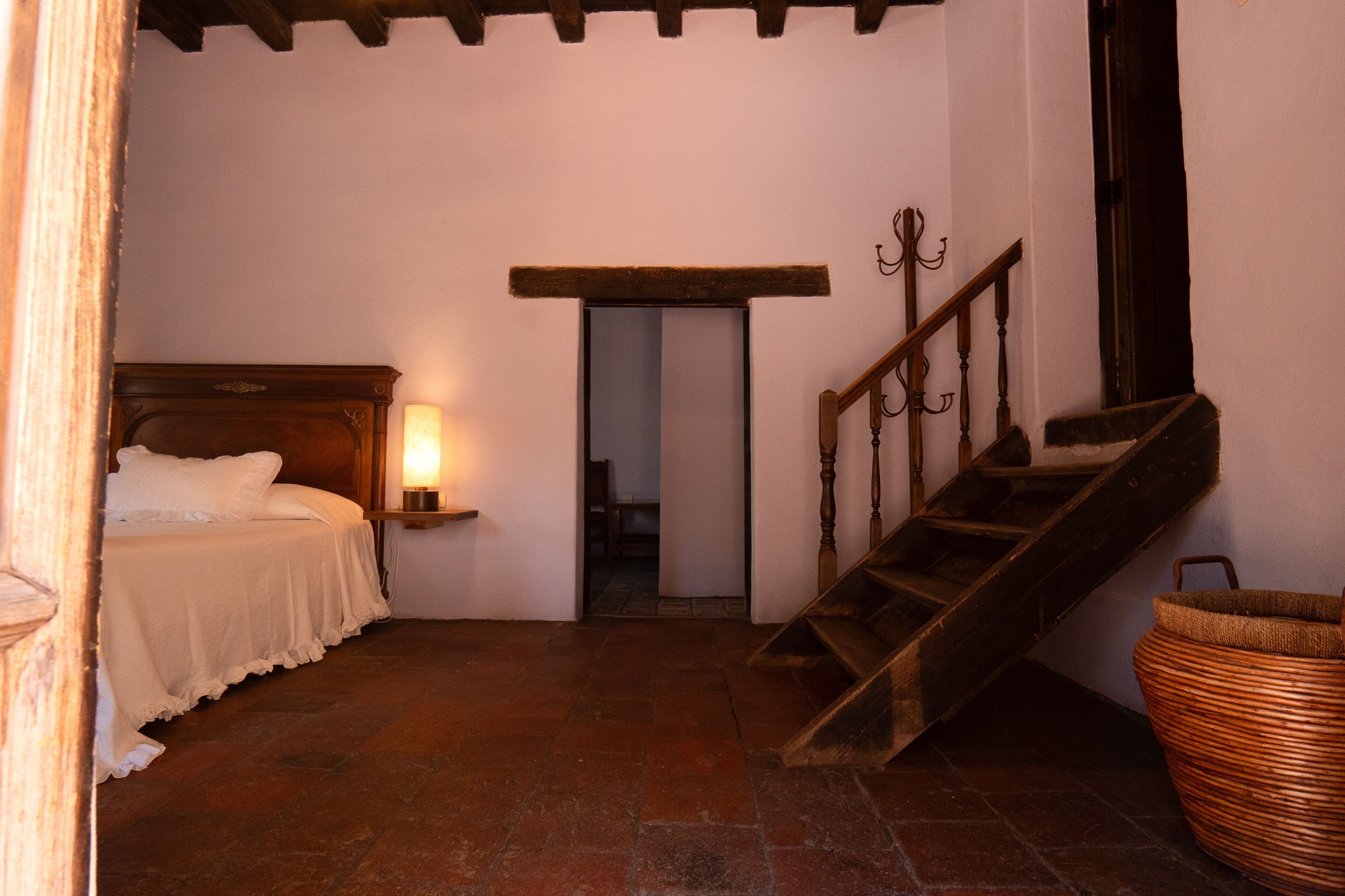 A bedroom with a bed, a nightstand with a lamp, a wooden staircase, and a woven basket.