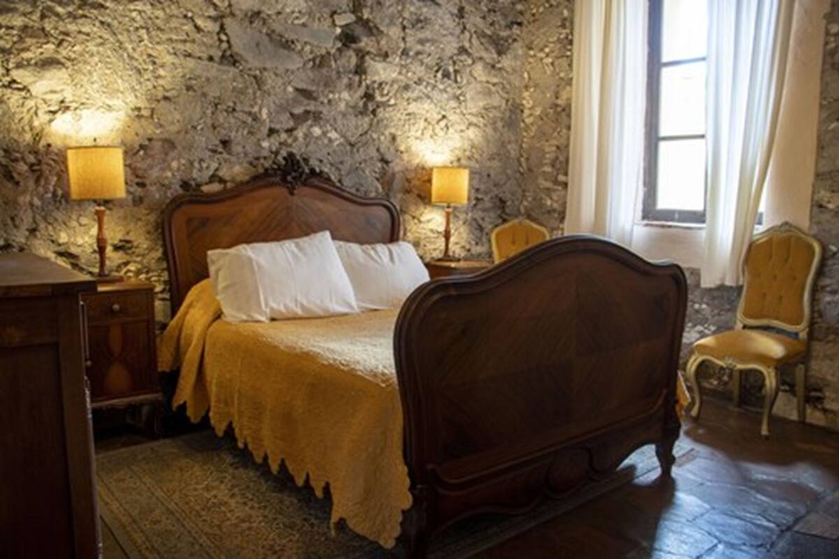 Une chambre avec un mur en pierre, un lit en bois, deux lampes de chevet, une chaise et une fenêtre avec des rideaux.