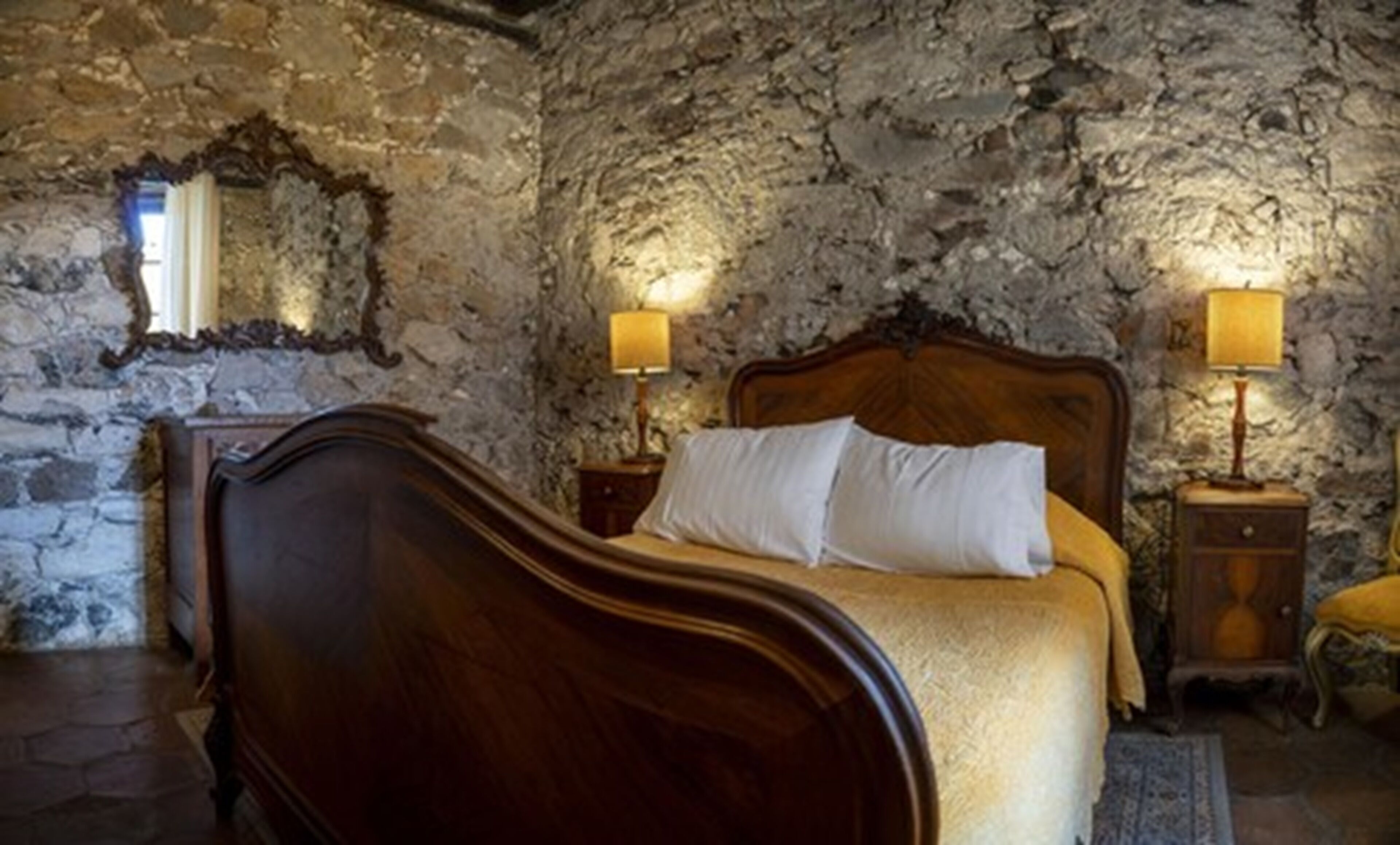 A bedroom with a stone wall, a wooden bed with white bedding, and two bedside lamps.