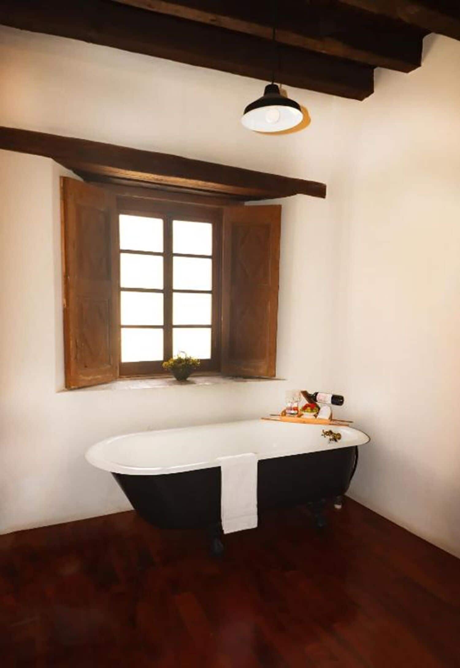 Un baño en esquina con una bañera independiente, una ventana con contraventanas de madera y una lámpara colgante.