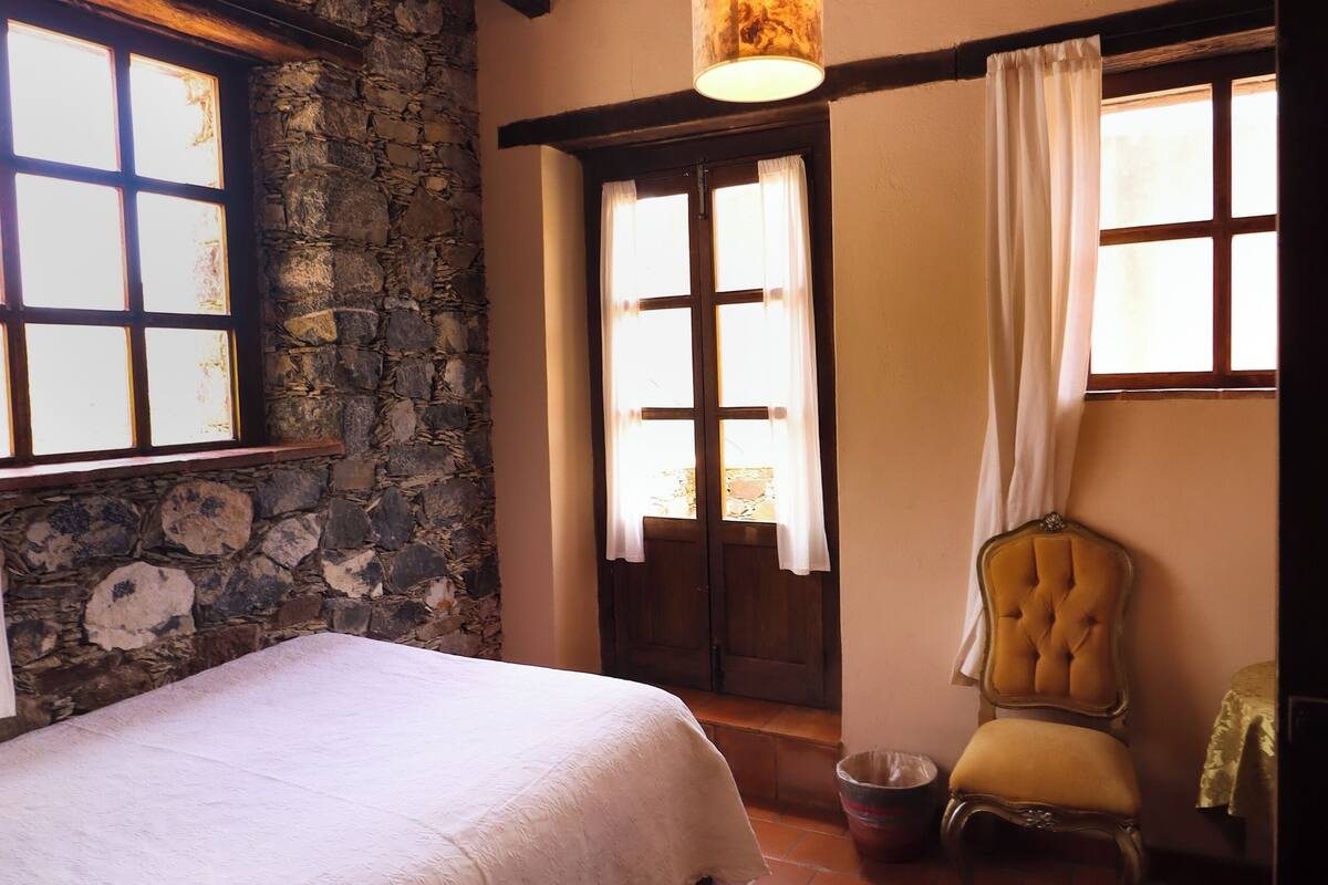 Une chambre avec un mur en pierre, des poutres apparentes, un lit recouvert d’une courtepointe blanche, un luminaire suspendu et une chaise.