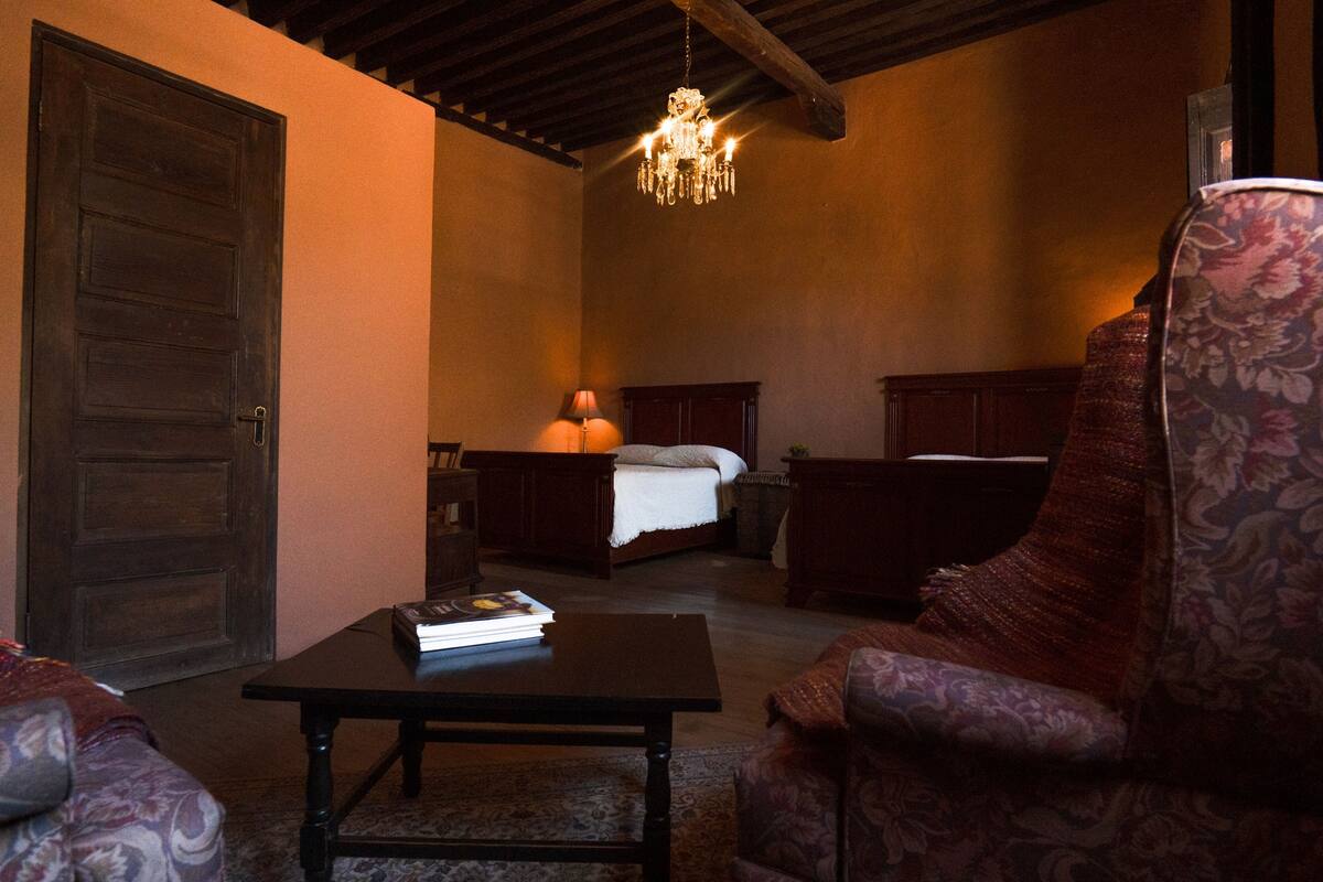 Une pièce avec une porte en bois, un lit recouvert d’une courtepointe blanche, une table de chevet, une chaise à motifs floraux et un lustre.