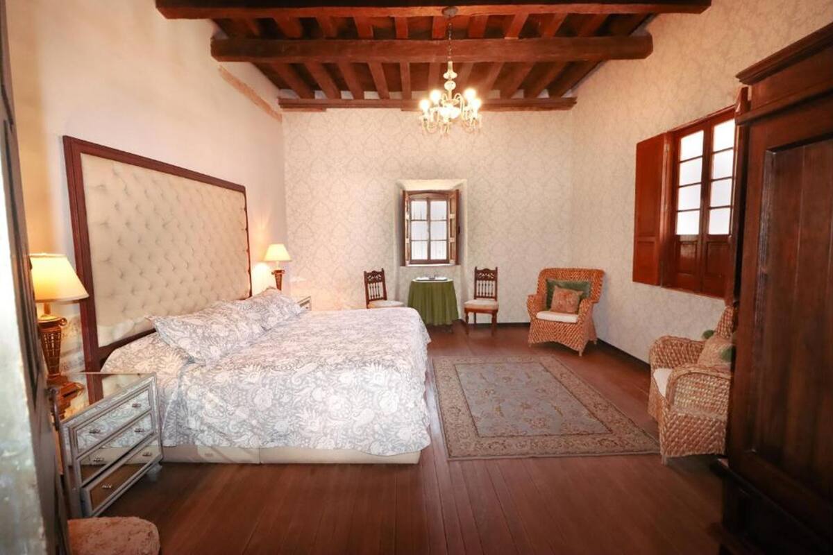 Une chambre à coucher avec un grand lit, deux fauteuils, une petite table, un lustre et des poutres en bois au plafond.