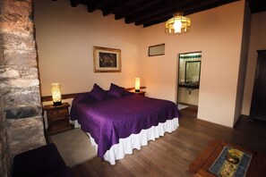 A bedroom with a large bed, two bedside lamps, a wooden nightstand, a stone wall, a framed picture on the wall, and a mirror in the corner. - Hotel Boutique Meson de la Abundancia (Real de Catorce)