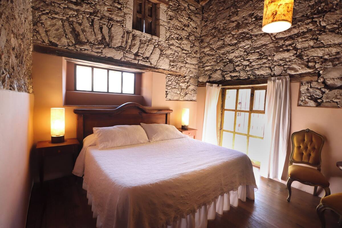Une chambre avec un plafond en pierre, un lit en bois, deux lampes de chevet, une chaise et une fenêtre avec des rideaux.