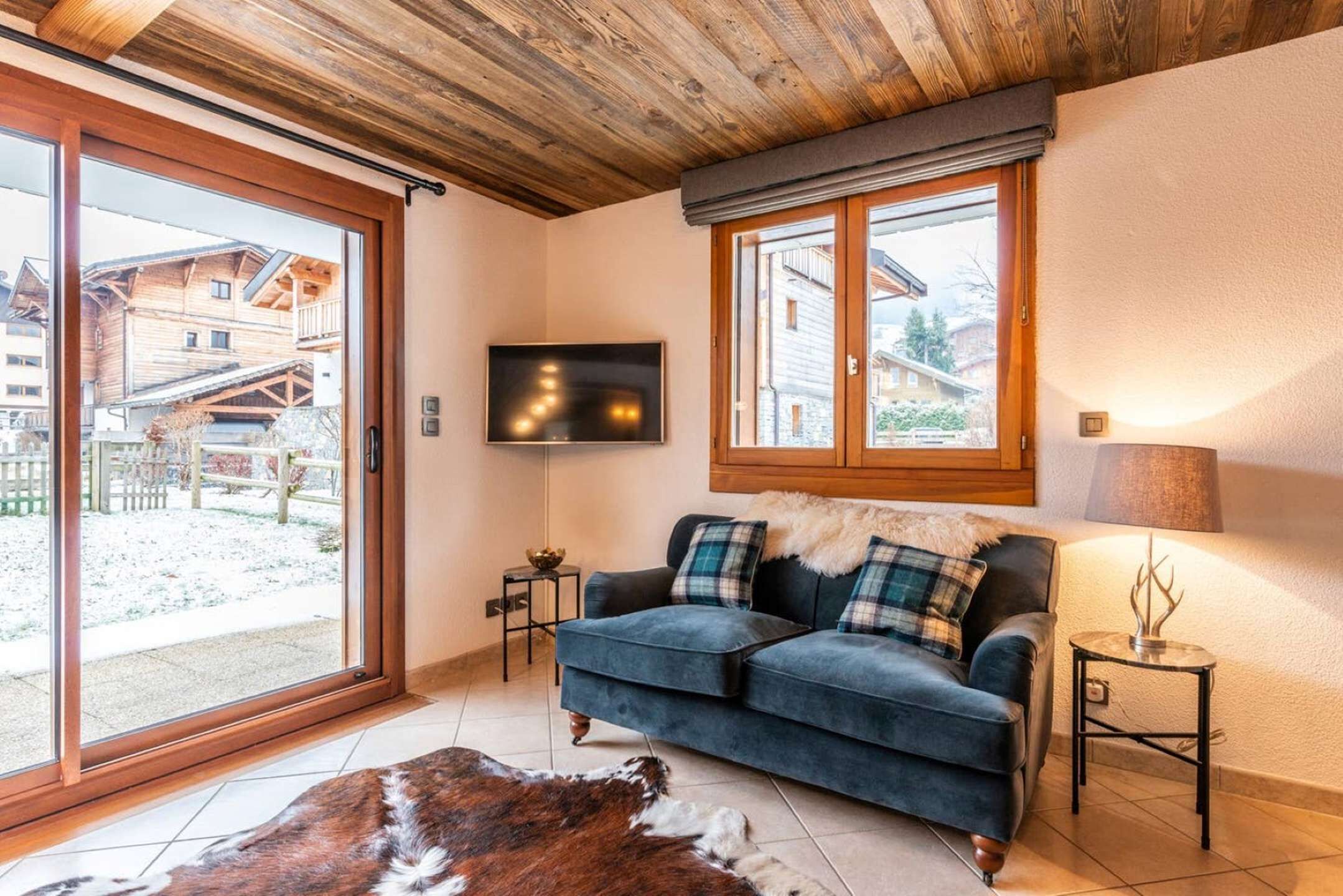 A cozy living room with a blue sofa, a wooden ceiling, a TV mounted on the wall, and a large window offering a view of a wooden building outside.