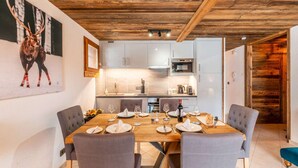 Un comedor con una mesa de madera preparada para cuatro personas, sillas grises tapizadas, y una cocina al fondo con armarios blancos y techo de madera.