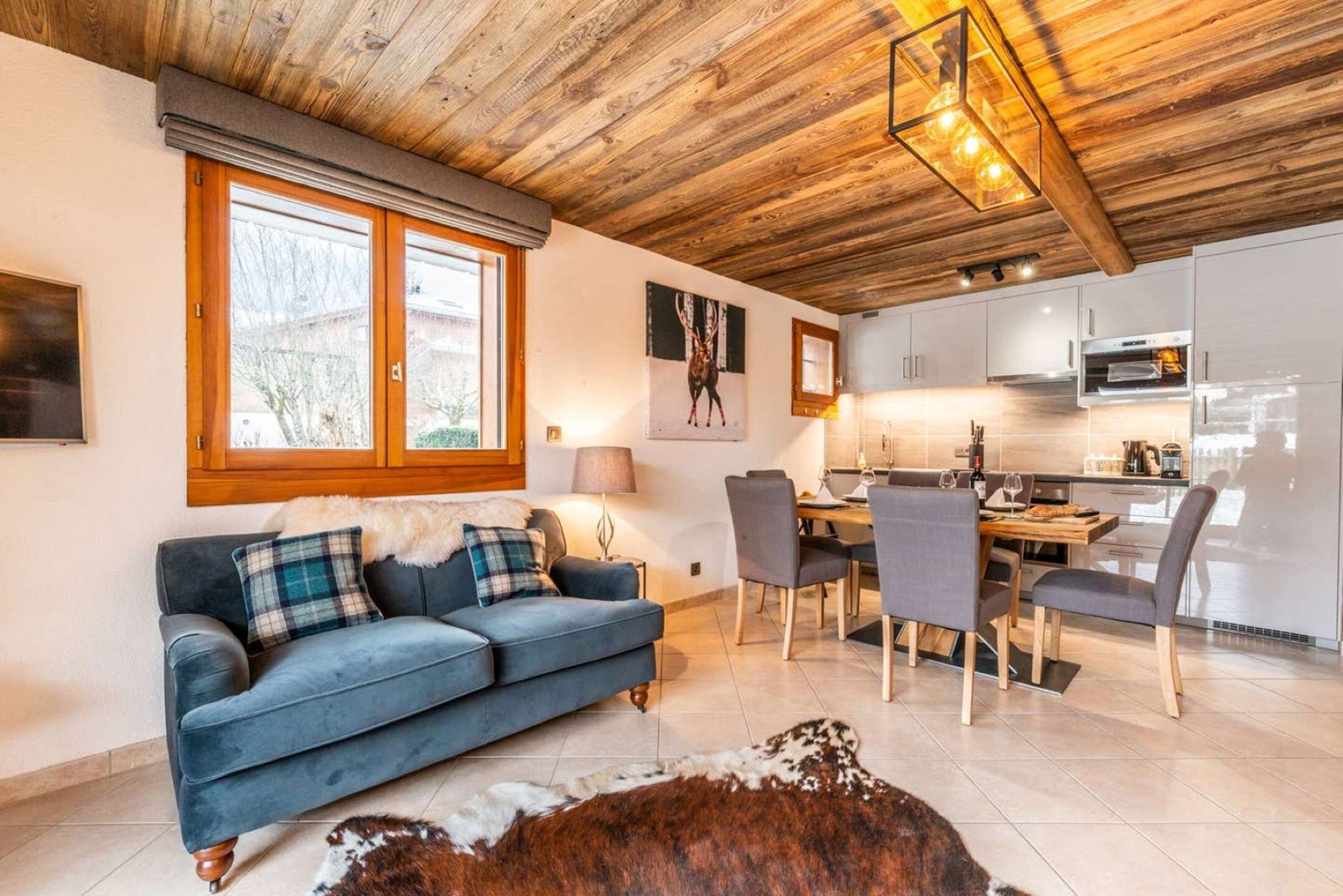Ett modernt vardagsrum med en blå soffa, en matplats med stolar, ett kök med vita skåp och ett träpanelat tak.