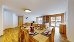 Ein Essbereich mit einem Holztisch, gedeckt für sechs Personen, umgeben von Holzstühlen. Ein Fernseher ist an der Wand befestigt, und rechts befindet sich ein Holzschrank.
