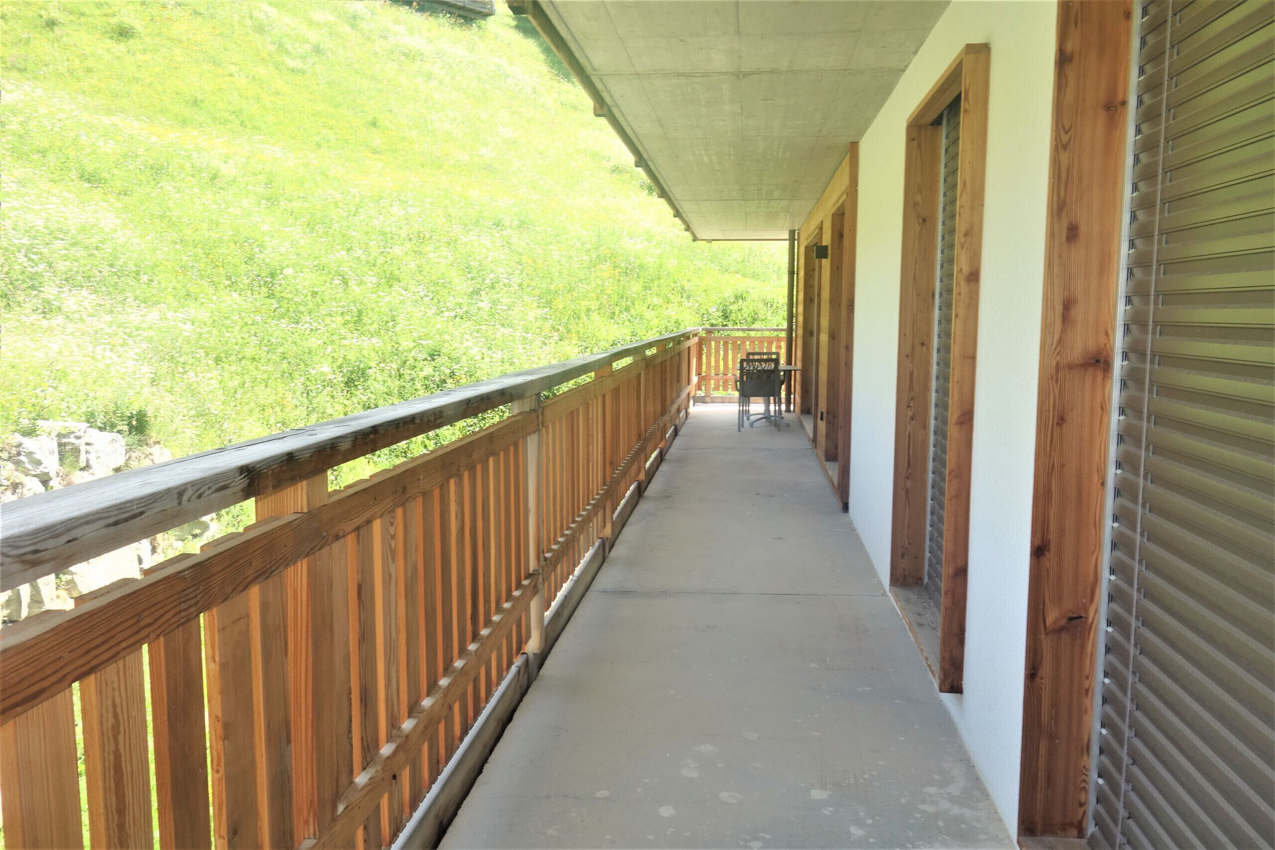 Un balcon abrité, doté d’une balustrade en bois, d’un sol en béton et offrant une vue sur une colline verdoyante.