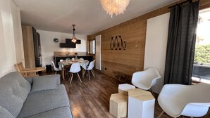 Un soggiorno moderno con un divano grigio, sedie bianche, un tavolo da pranzo per quattro persone, una cucina sullo sfondo, pareti in legno e un'ampia finestra con tende.