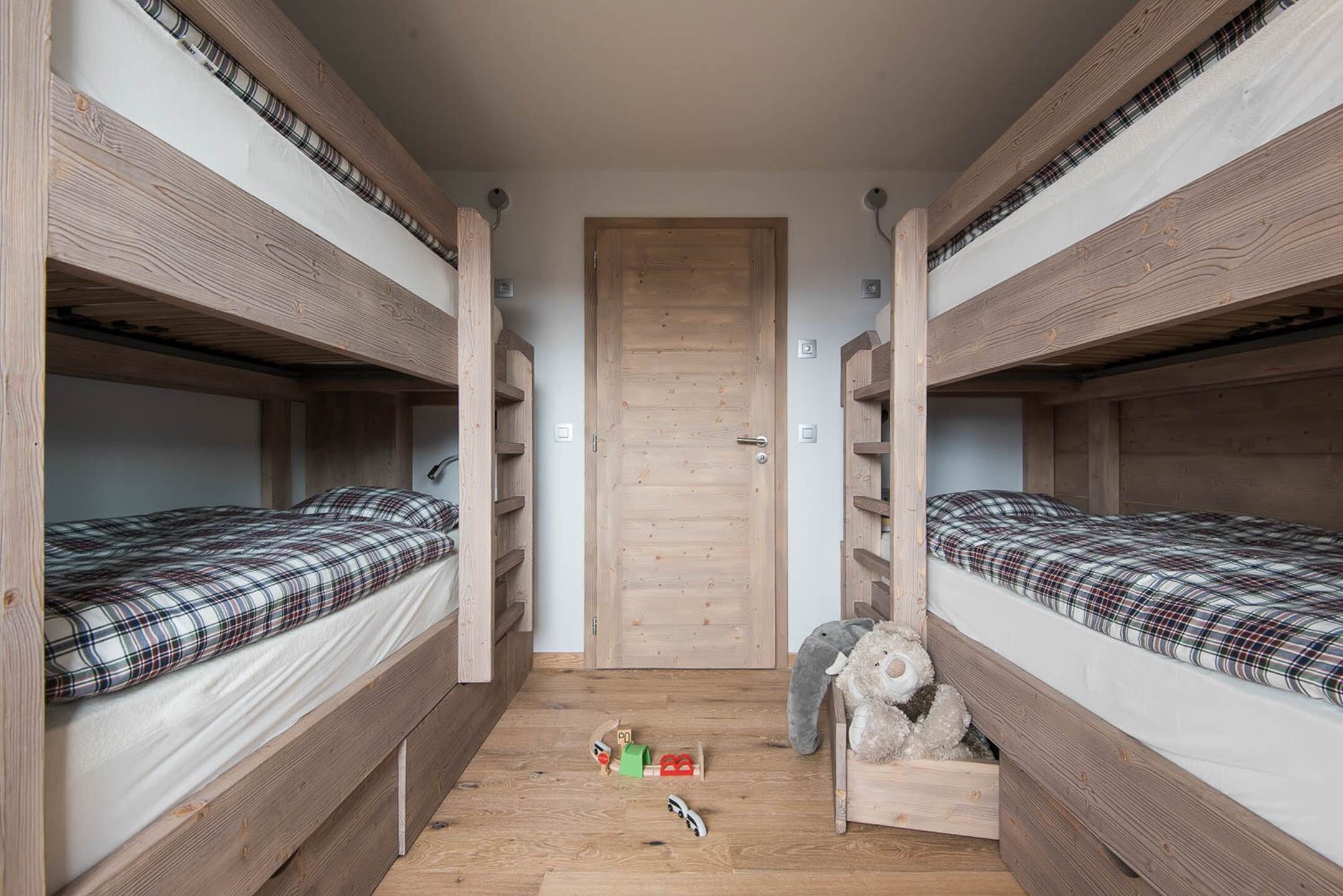 A bunk bed room with a wooden door, two beds with plaid bedding, and a teddy bear on the right bed.